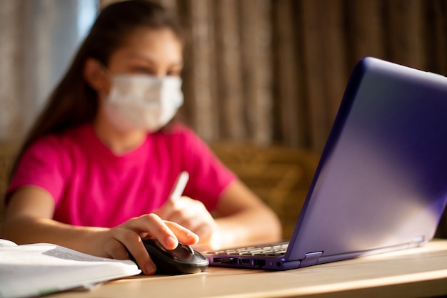 Child wearing face medical mask self-studying at home during coronavirus outbreak. Young schoolgirl having distance learning works at the computer