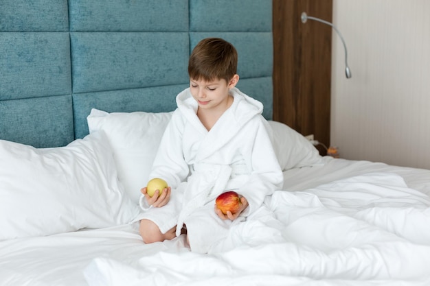 Un bambino in camice bianco sul letto sorride e tiene in mano mele health morning