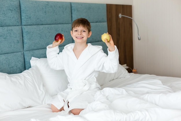 Un bambino in camice bianco sul letto sorride e tiene in mano mele health morning