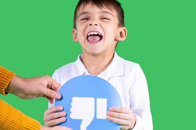 Child in white shirt is holding in his arms a paper circle form blue color with a sign thumb down green background