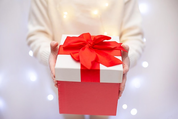 Child in white sweater holding red christmas gift in hand on light bokeh background