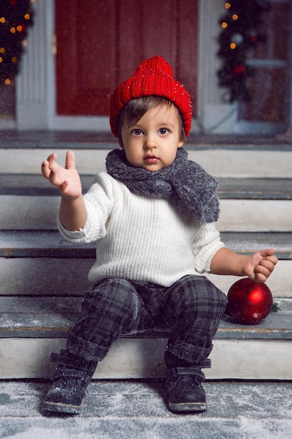 Photo child in a white sweater and a red knitted hat sits on the steps with snow at home at christmas