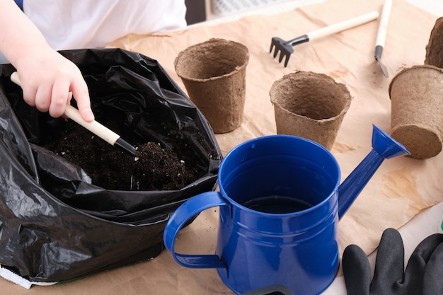白いTシャツを着た子供が泥炭の種の鉢に土を注ぎ、子供がテーブルの上にセネン、園芸工具を植えます