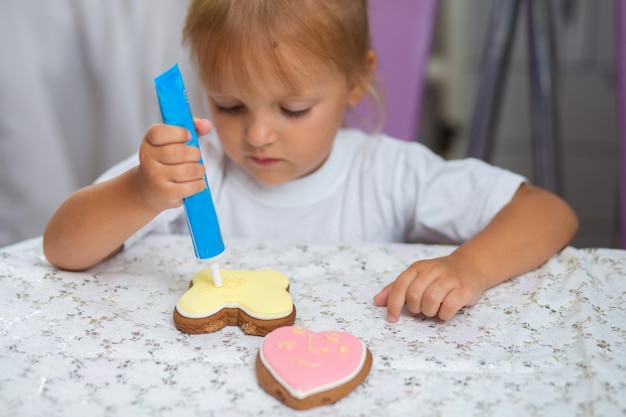 The child in a white t-shirt sits at a table and draws cream on cookies