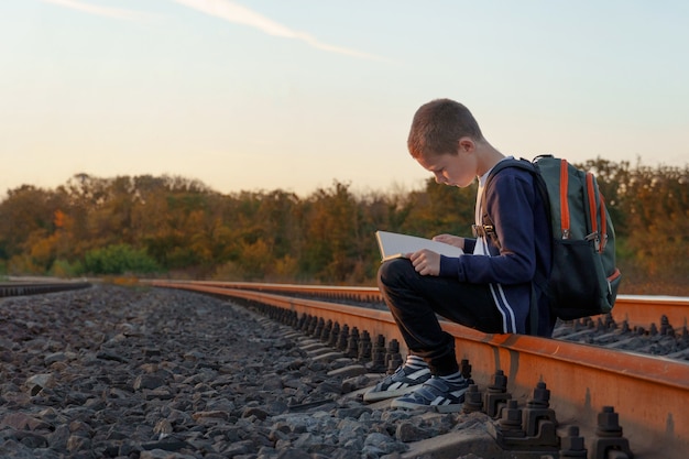 Фото Ребенок с книгой в руках на железнодорожных рейсах. мальчик сидит на рейсах и читает книгу на фоне заходящего солнца.