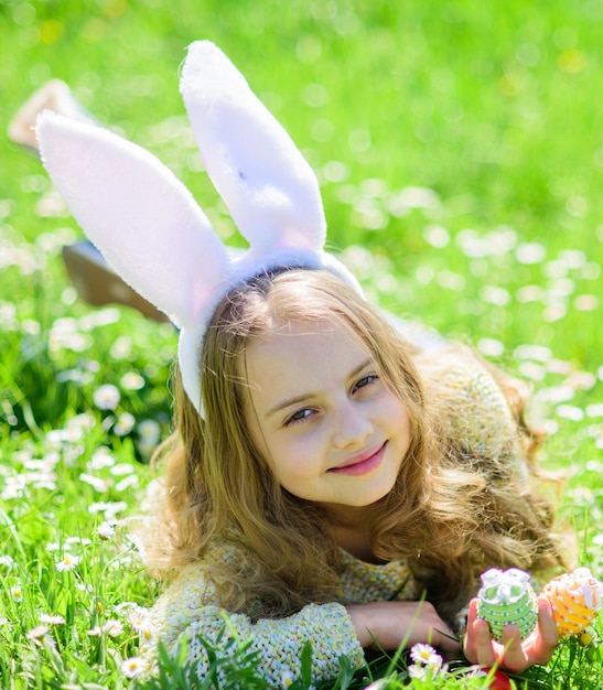 Child with cute bunny ears lying at meadow. Little girl hunting for Easter egg in spring garden on Easter day. Cute child lay on grass with eggs in hands. Traditional bunny celebrating feast concept.