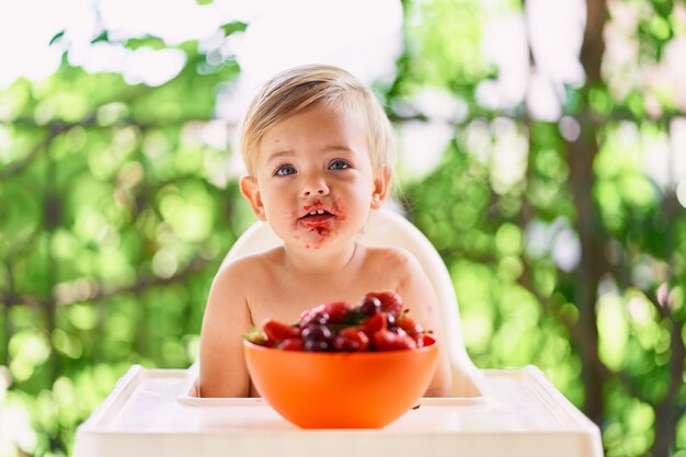 汚れた顔の子供は果物のプレートの前のテーブルに座っています