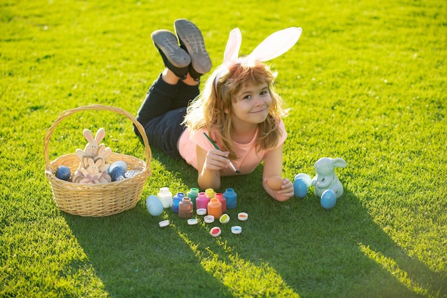 Child with easter eggs and bunny ears laying on grass painting eggs. Happy Easter kids face.