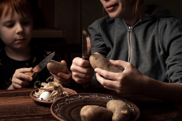 Child with mom peels potatoes on a dark background A woman teaches her son to peel potatoes