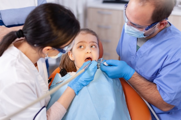 Child with mouth open in the course of caries treatment looking at dentist. Mother with her kid in stomatology clinic for teeth examine using modern instruments.