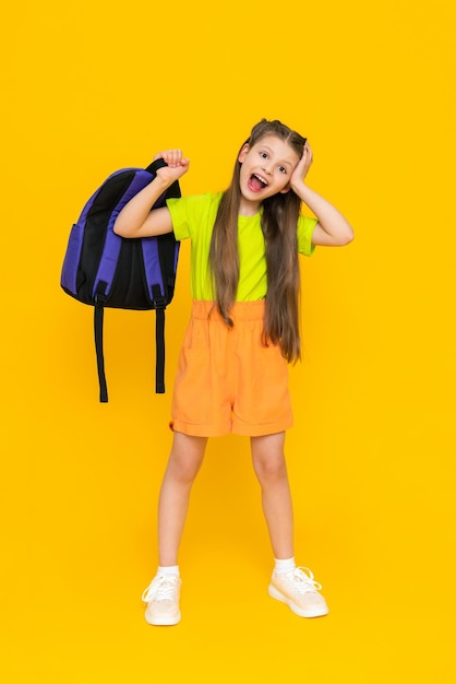 A child with a school backpack A beautiful little girl in full height in shorts and a Tshirt Education during the summer holidays Preparatory courses for children Yellow isolated background