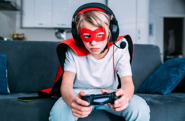 Bambino con costume da super eroe che gioca a un videogioco con joystick seduto sul divano di casa - concetto di bambini, giochi e tecnologia