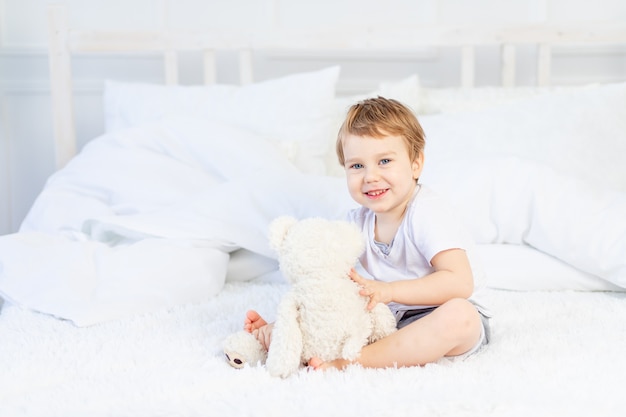 A child with a teddy bear on the bed at home plays and laughs