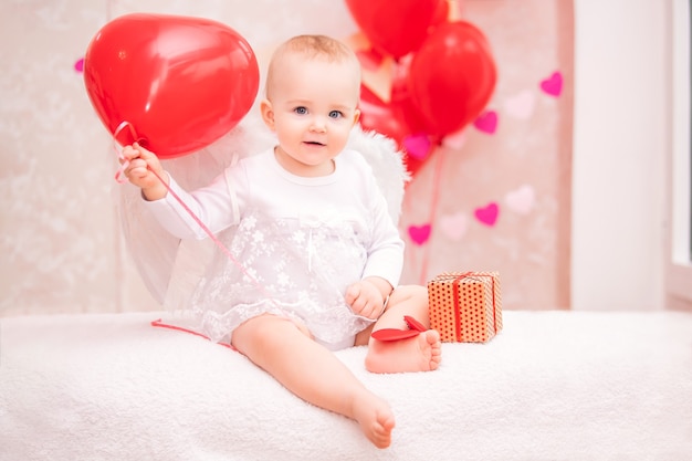 Ребенок с белыми крыльями из перьев держит красный воздушный шар в форме сердца, символ Дня святого Валентина.