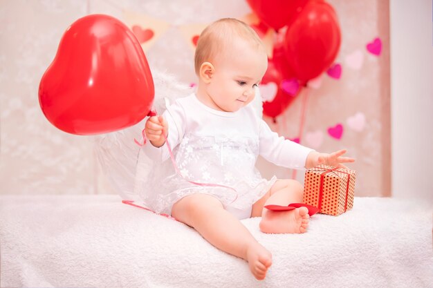 Child with white feathered wings holding a red balloon in the shape of a heart and sees a box with a gift,a symbol of Valentine's Day.