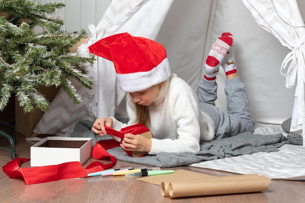 クリスマスのサプライズ プレゼントをひもでクラフト紙に包む子供 サンタの帽子をかぶった女の子が自宅で家族へのプレゼントを詰めている