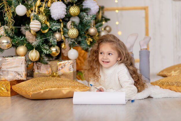 A child writes a letter for Santa Claus under a gold-decorated Christmas tree