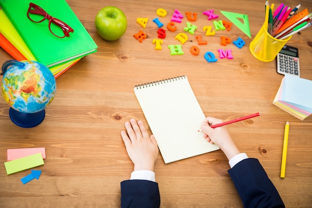 Child writing in copybook. School items on wooden desk in class. Education concept. Top view