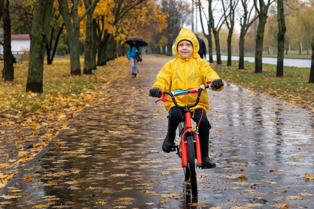 Child in yellow raincoat rides bicycle in an autumn park. Boy on bicycle in the rain. Portrait of happy preschooler.