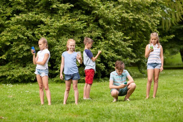 子供の頃、拡張現実、インターネット中毒、テクノロジー、人のコンセプト – 夏の公園でスマートフォンを持った子供や友人のグループ