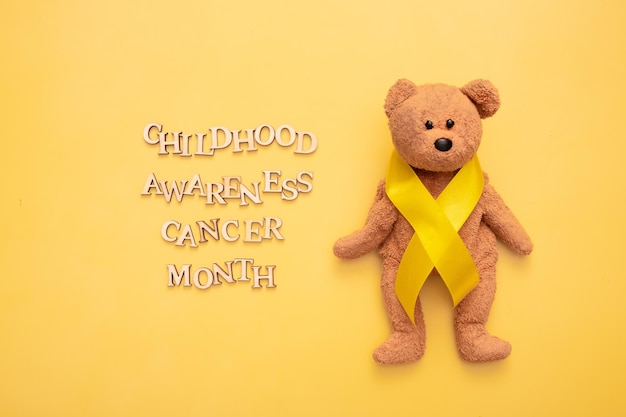 Photo childhood cancer awareness month text from wooden letters and teddy bear with yellow ribbon on yellow background