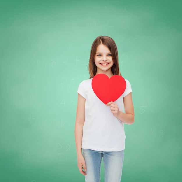 childhood, charity, education, love and people concept - smiling little girl sitting with red heart cutout over green blackboard background