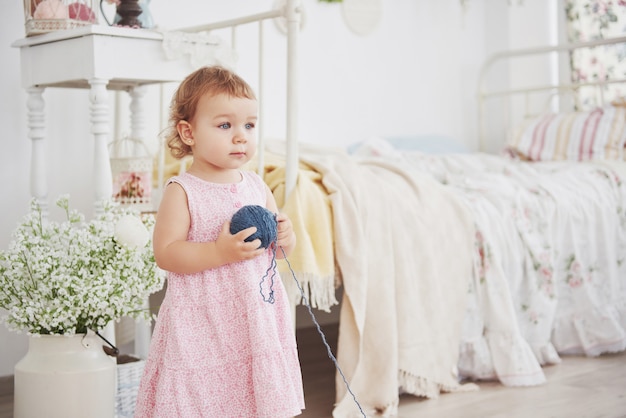 Childhood concept. Baby girl in cute dress play with colored thread. White vintage childroom