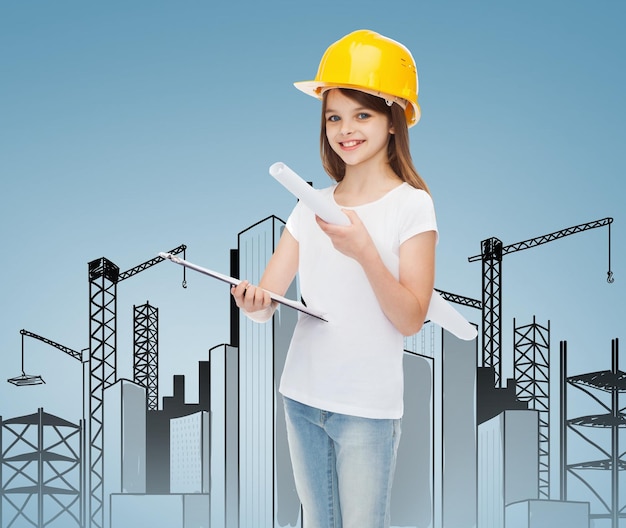 childhood, construction, architecture, building and people concept - smiling little girl in protective helmet with clipboard and blueprint