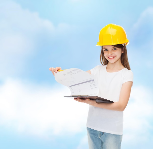 childhood, construction, architecture, building and people concept - smiling little girl in protective helmet with clipboard turning page