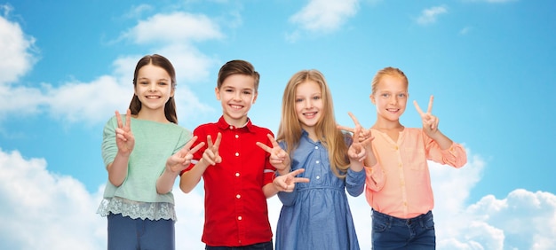 childhood, fashion, friendship and people concept - happy smiling boy and girls showing peace hand sign over blue sky and clouds background