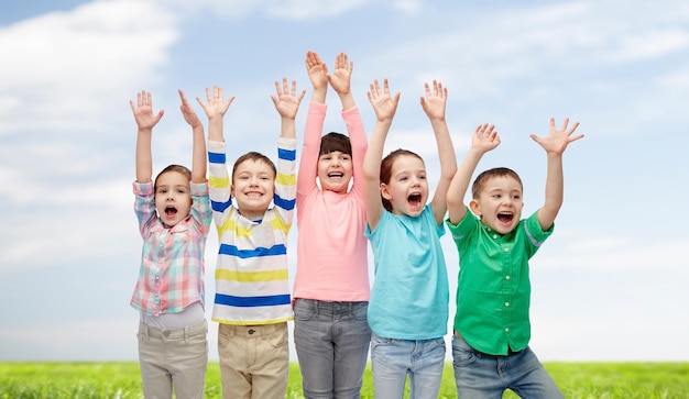 childhood, fashion, gesture and people concept - happy smiling friends raising fists and celebrating victory over blue sky and grass background