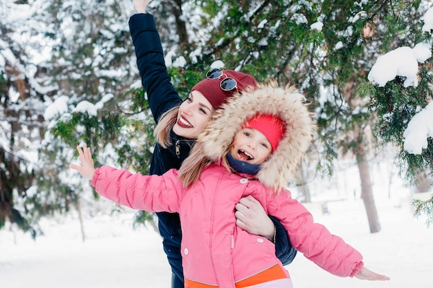 Infanzia, moda, stagione e concetto di persone - cute little girl guardando sua madre che sta spazzando via la neve. famiglia felice durante la passeggiata invernale all'aperto al mattino con tempo freddo e soleggiato.