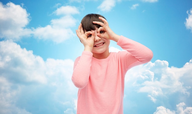 childhood, fun, gesture and people concept - happy little girl making faces over blue sky and clouds background