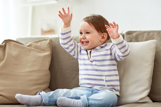 childhood, fun and people concept - happy smiling baby girl sitting on sofa at home