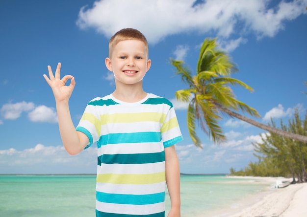 Concetto di infanzia, gesto, estate, viaggi e persone - ragazzino sorridente che fa segno ok su sfondo spiaggia tropicale