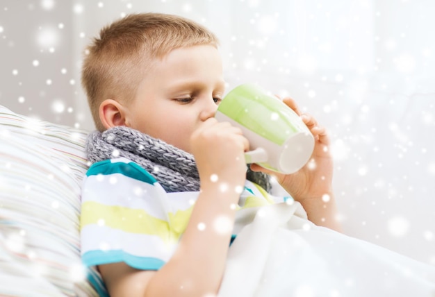 childhood, healthcare and people concept - ill boy with flu in bed drinking from cup at home