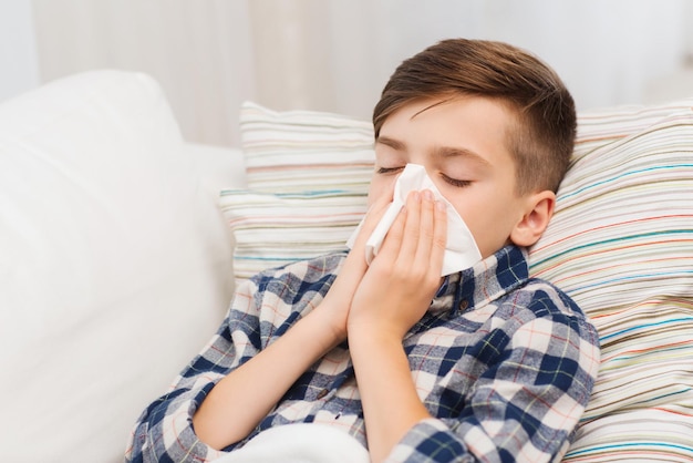 childhood, healthcare, rhinitis, people and medicine concept - ill boy with flu lying in bed and blowing his nose at home