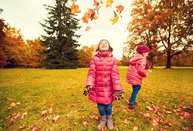 childhood, leisure, friendship and people concept - group of happy little girl playing with autumn maple leaves and having fun in park