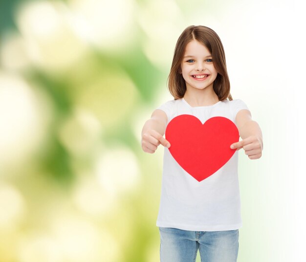 childhood, love, charity, ecology and people concept - smiling little girl sitting and holding red heart cutout over green background