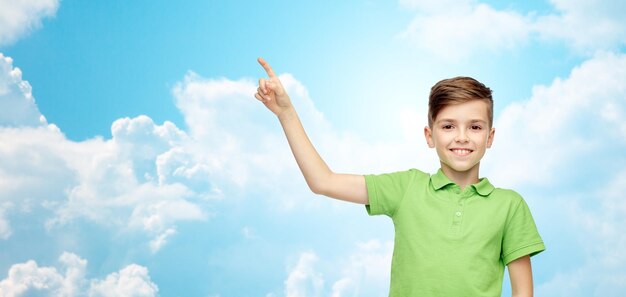 Childhood and people concept - happy smiling boy in green polo t-shirt pointing finger up over blue sky and clouds background