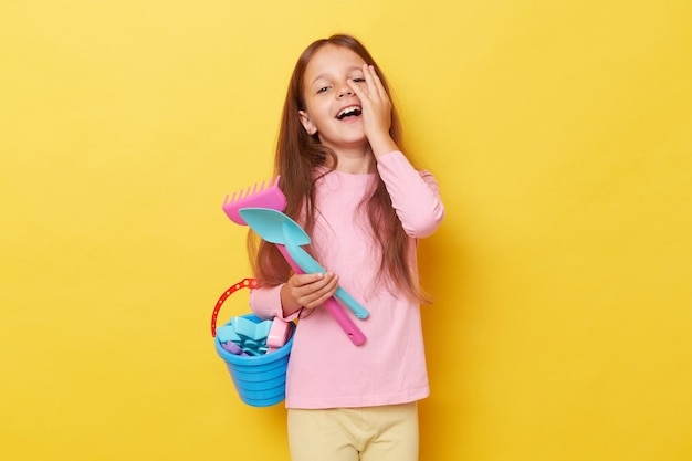 Childhood sandbox joy Springtime play kit Happy funny little cute girl with dark hair holding beach sandbox toys rake and shovel isolated over yellow background laughing while having time