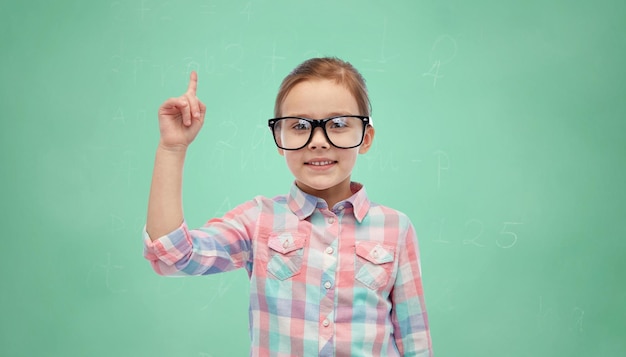 childhood, school, education, vision and people concept - happy little girl in eyeglasses pointing finger up over green school chalk board background