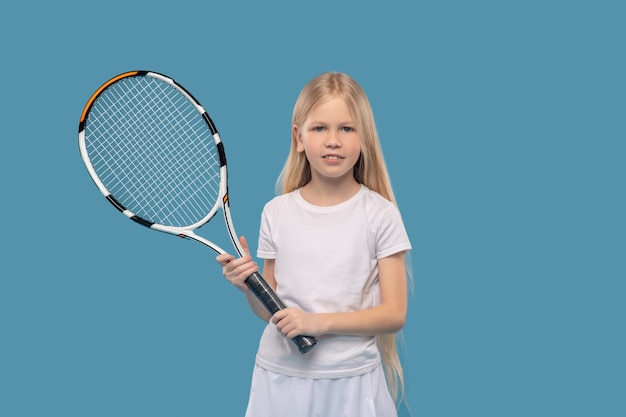 Childhood, sports. Happy caucasian elementary school girl in white tshirt with professional tennis racket on blue background