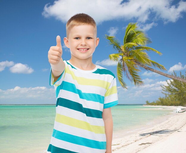 어린 시절, 여름 방학, 여행, 몸짓, 그리고 사람들의 개념 - 열대 해변 배경 위에 엄지손가락을 치켜드는 웃는 어린 소년