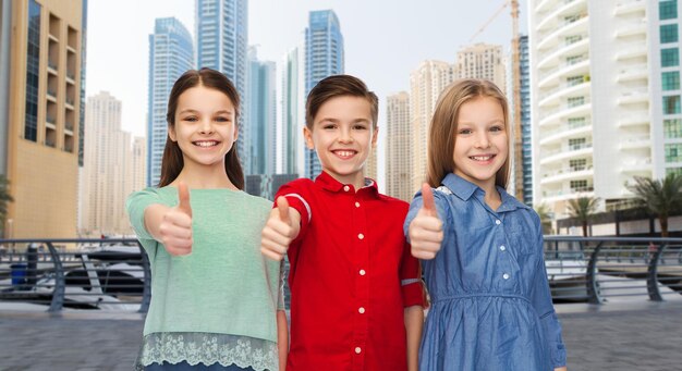 childhood, travel, tourism, gesture and people concept - happy smiling boy and girls showing thumbs up over dubai city street background