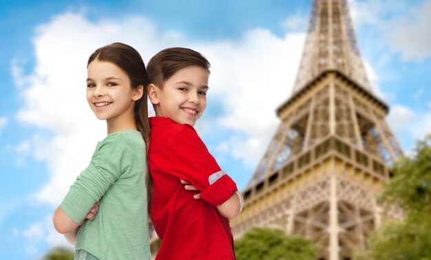 Childhood, travel, tourism, and people concept - happy smiling boy and girl standing back to back over paris eiffel tower background