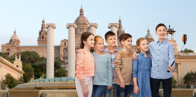 childhood, travel, tourism, technology and people concept - happy children talking picture by smartphone on selfie stick ovetrnational museum of barcelona background