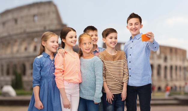 childhood, travel, tourism, technology and people concept - happy children talking selfie by smartphone over coliseum in rome