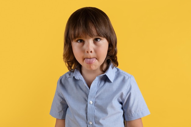 Photo childish pampering cute little boy fooling around sticking his tongue out and squinting his eyes orange background