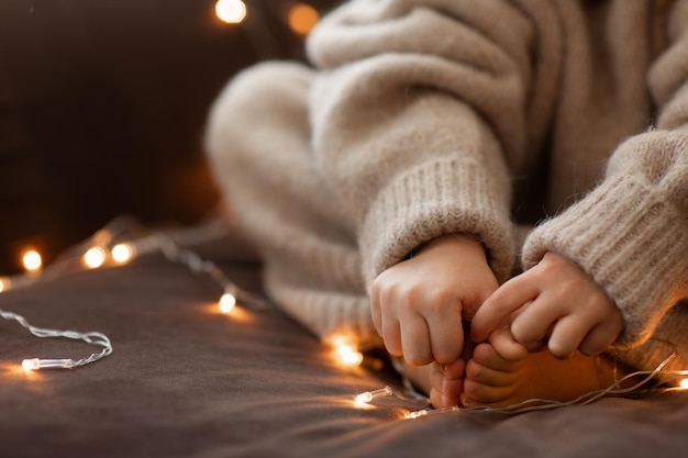 Children bare feet and hands close up garland lights. flaffy\
fuzzy warm knitted beige sweater. christmas concept, holiday.happy\
new year.selective focus. child girl sitting on sofa inside.cozy\
vibes